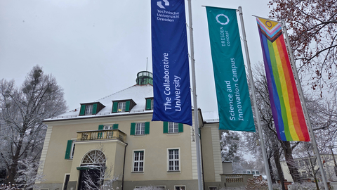 Three flags—TU Dresden, Dresden Concept, and a Pride flag—wave in front of a snowy university building. Trees and surroundings are covered in winter snow.
