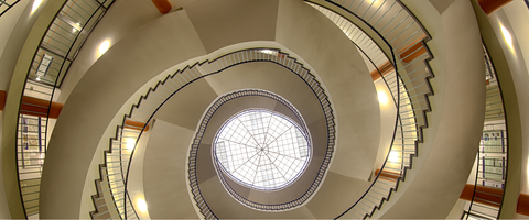 Treppenaufgang im Hülße-Bau der TU Dresden. Das Bild wurde von unten aufgenommen. Den Mittelpunkt bildet das Dachfenster, dessen Konstruktion wie ein Spinnennetz aussieht. Von unten betrachtet läuft die Treppe spiralförmig nach oben. 